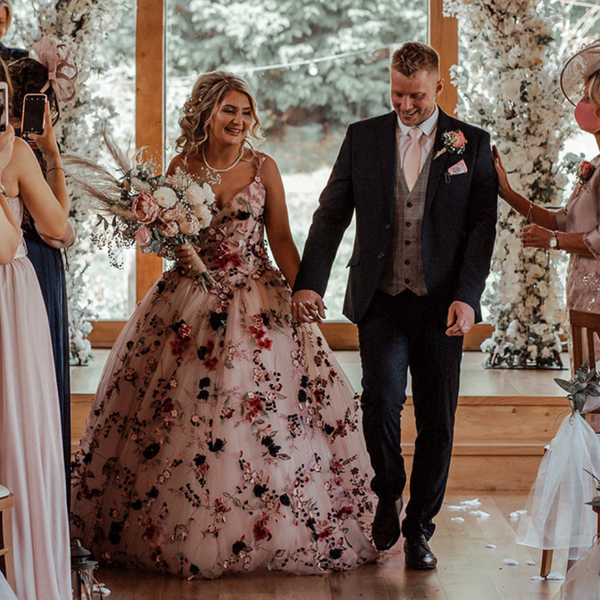 Ronald Joyce real bride, Rebecca, and her husband, Michael, holding hands on their wedding day. Rebecca has curled blonde hair and wears Celestina, our colourful ballgown wedding dress style with floral appliques. 